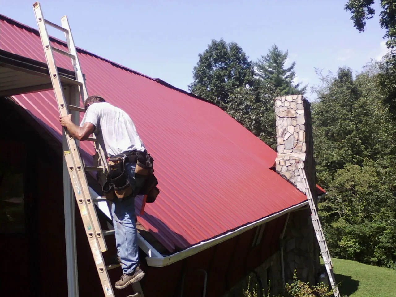 Roofer on a ladder beside a red metal roof and stone chimney — Soffit & Fascia Repair by Roofing Brothers in Round Mountain